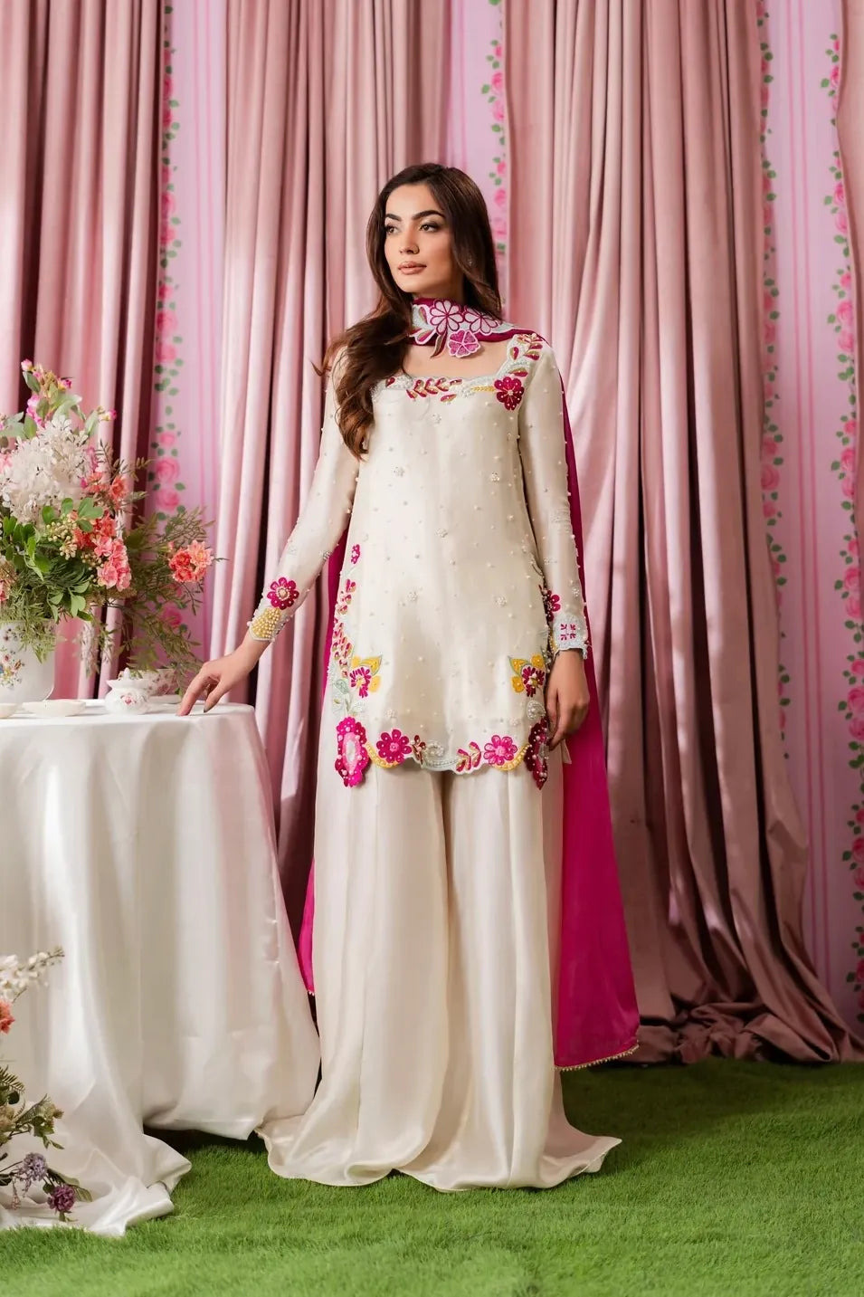 A direct frontal view of the Shanzay Anooshay Multi Floral outfit. The full scope of the cream Shamoz silk kurta, its rich embroidery, and the beautiful wide-leg satin trousers are showcased. The striking magenta dupatta trails dramatically behind her, emphasizing the flow of the fabric in the decorated indoor setting.