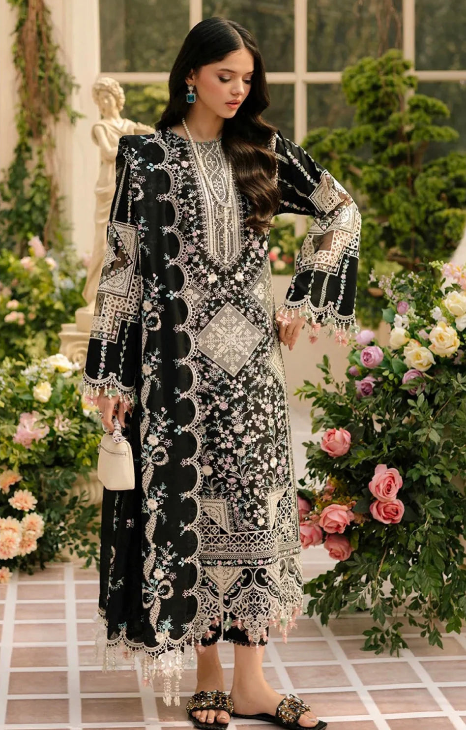Woman in an elegant black and white embroidered outfit standing in a decorative setting with flowers and a statue.