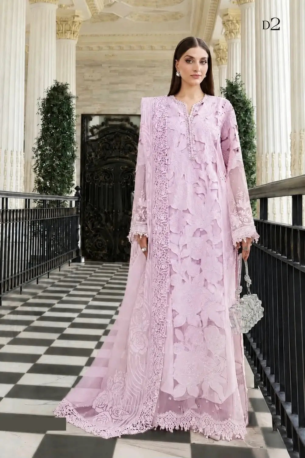 A fashion photograph, very similar to Image 6, showing the woman in the full-length, embroidered lilac suit standing and facing forward in the columned hallway. She is holding the embellished silver handbag, and the trailing dupatta is visible on the checkered floor.