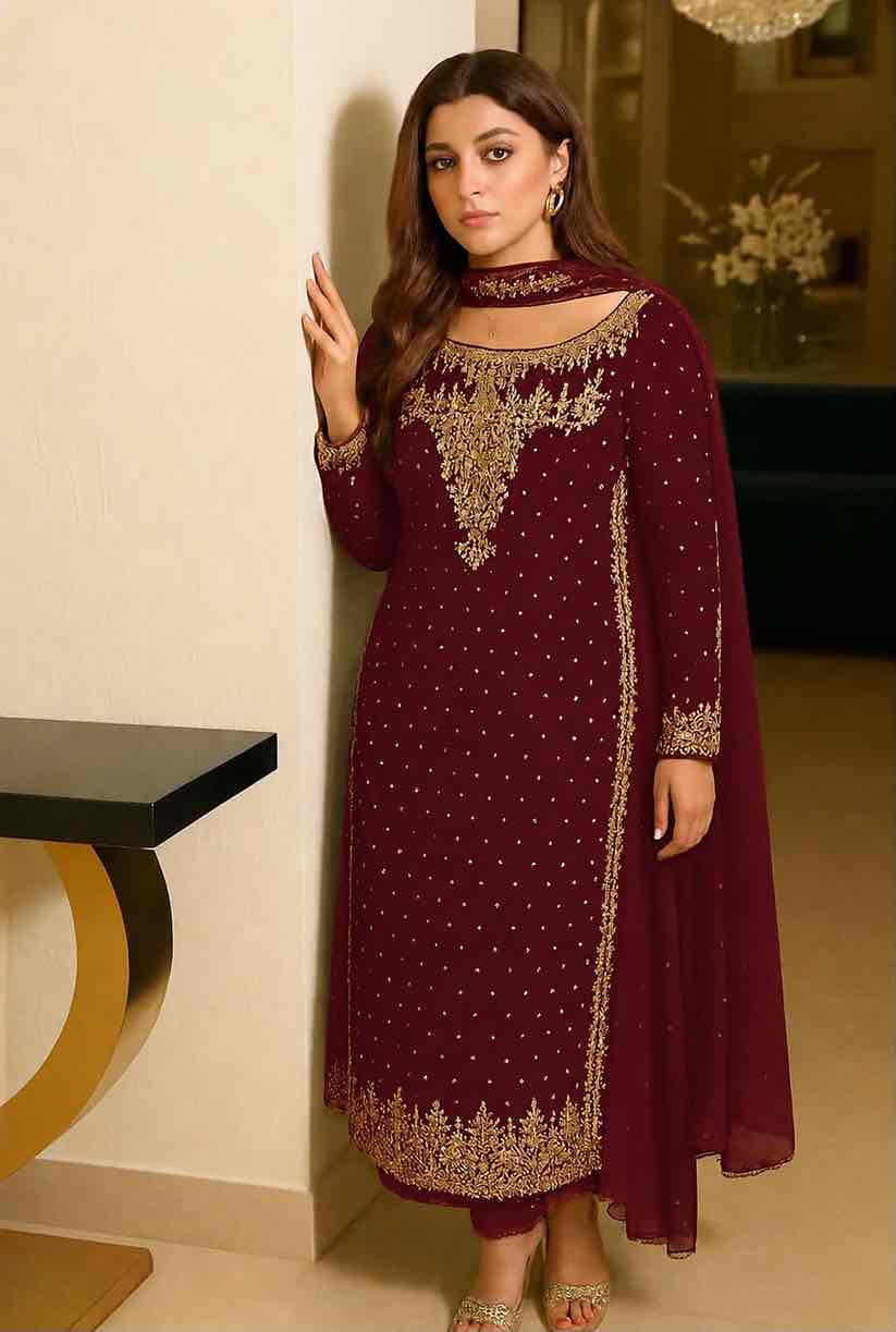 Model standing indoors against a neutral wall wearing long-sleeved maroon embellished chiffon dress with gold embroidered neckline and borders.
