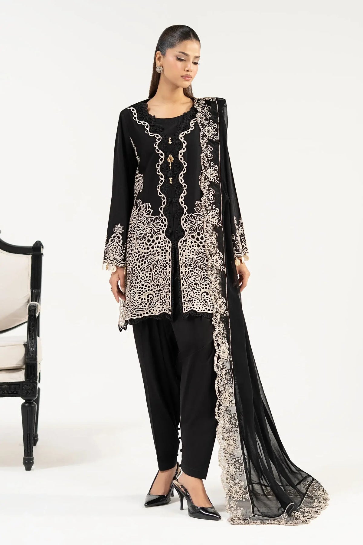 A full-length photograph of a woman standing and posing in a formal black Maria B three-piece suit. She is turned slightly towards the camera, showcasing the front of the long kurta with its heavy gold thread embroidery and scalloped edges. A classic black and cream armchair is partially visible to the side.
