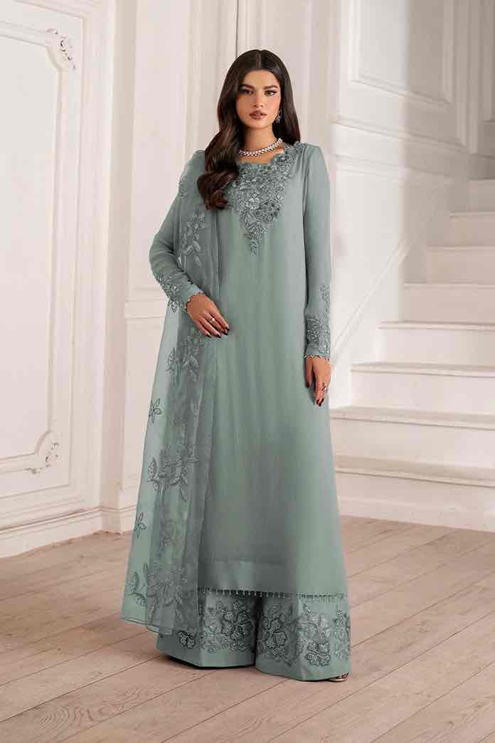 Full-length portrait of a woman standing and looking directly at the camera, wearing the Iznik Green Hand Embellished and embroidered Chiffon Dress. Her hands are elegantly placed at her sides. The dress has wide-leg trousers and is adorned with intricate silver floral embroidery. A matching embroidered dupatta drapes around her shoulder and arm. The background is a clean white classical interior with wainscoting. Shop Now.